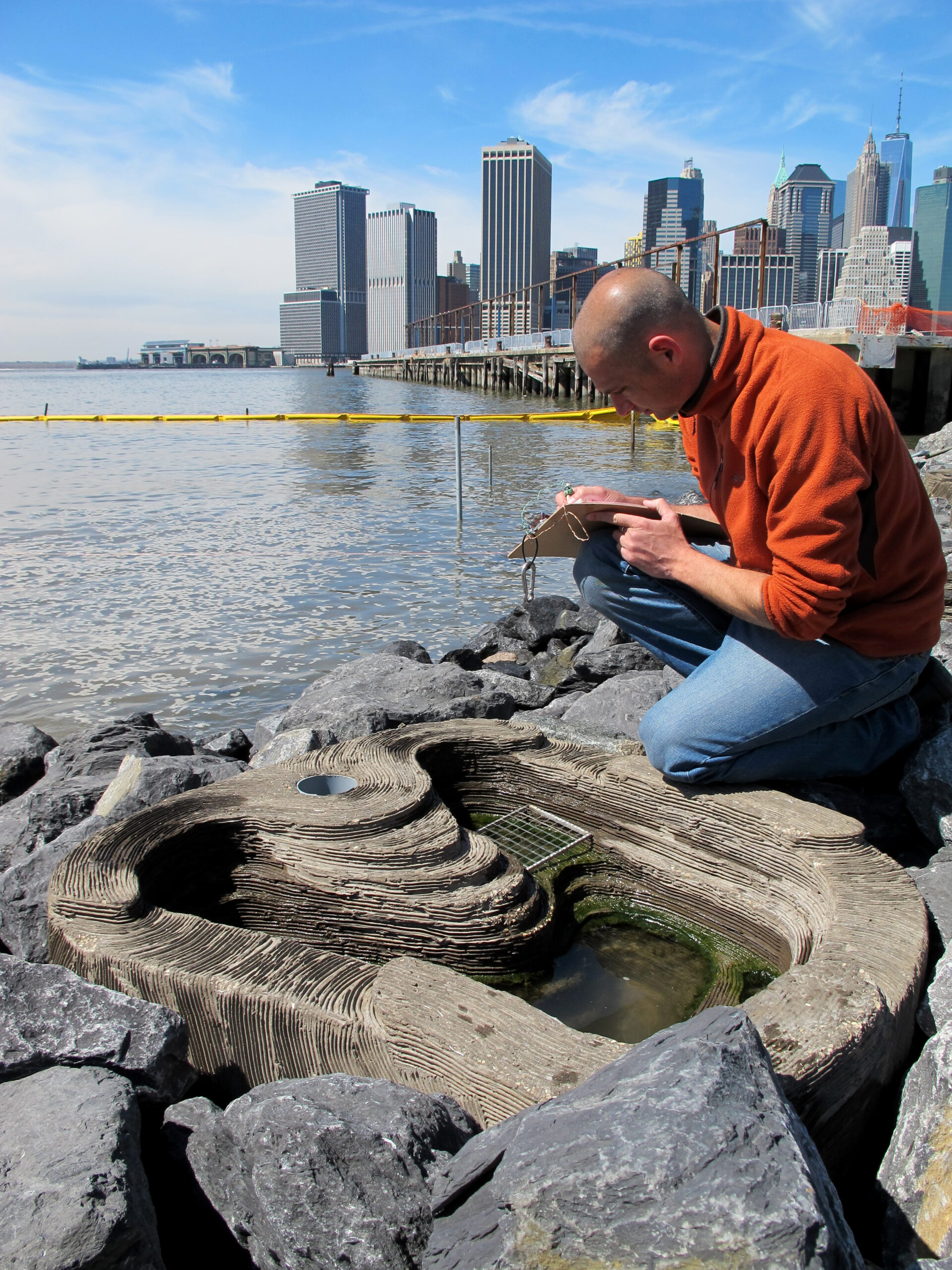 Balding man kneeling next to a tidepool on the coast, writing on a clipboard 